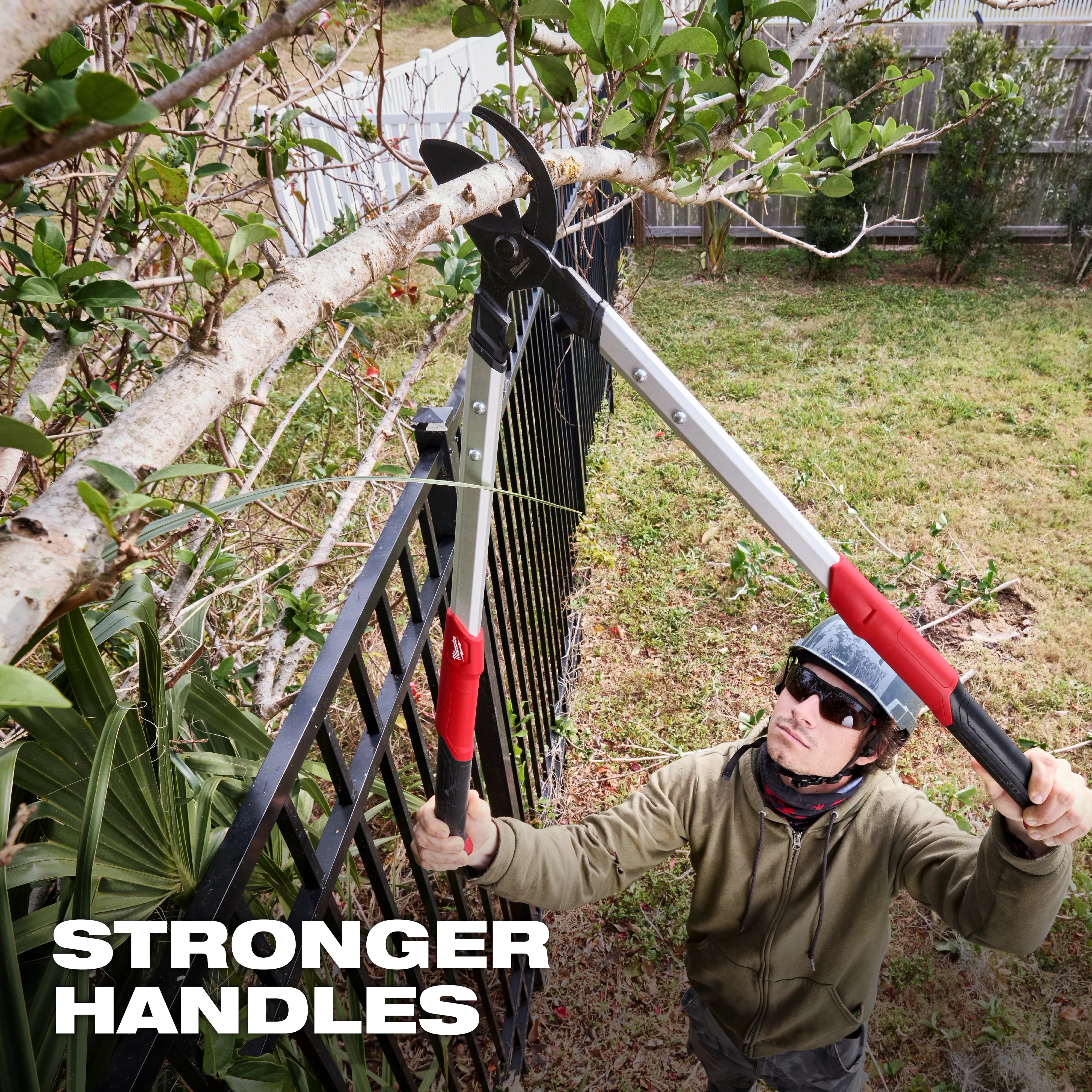 A person is using a 2" Lopper to cut a tree branch. The long-handled tool has red grips and is positioned over a fence. The image features text stating "STRONGER HANDLES" in bold white letters, emphasizing the durability of the handles.