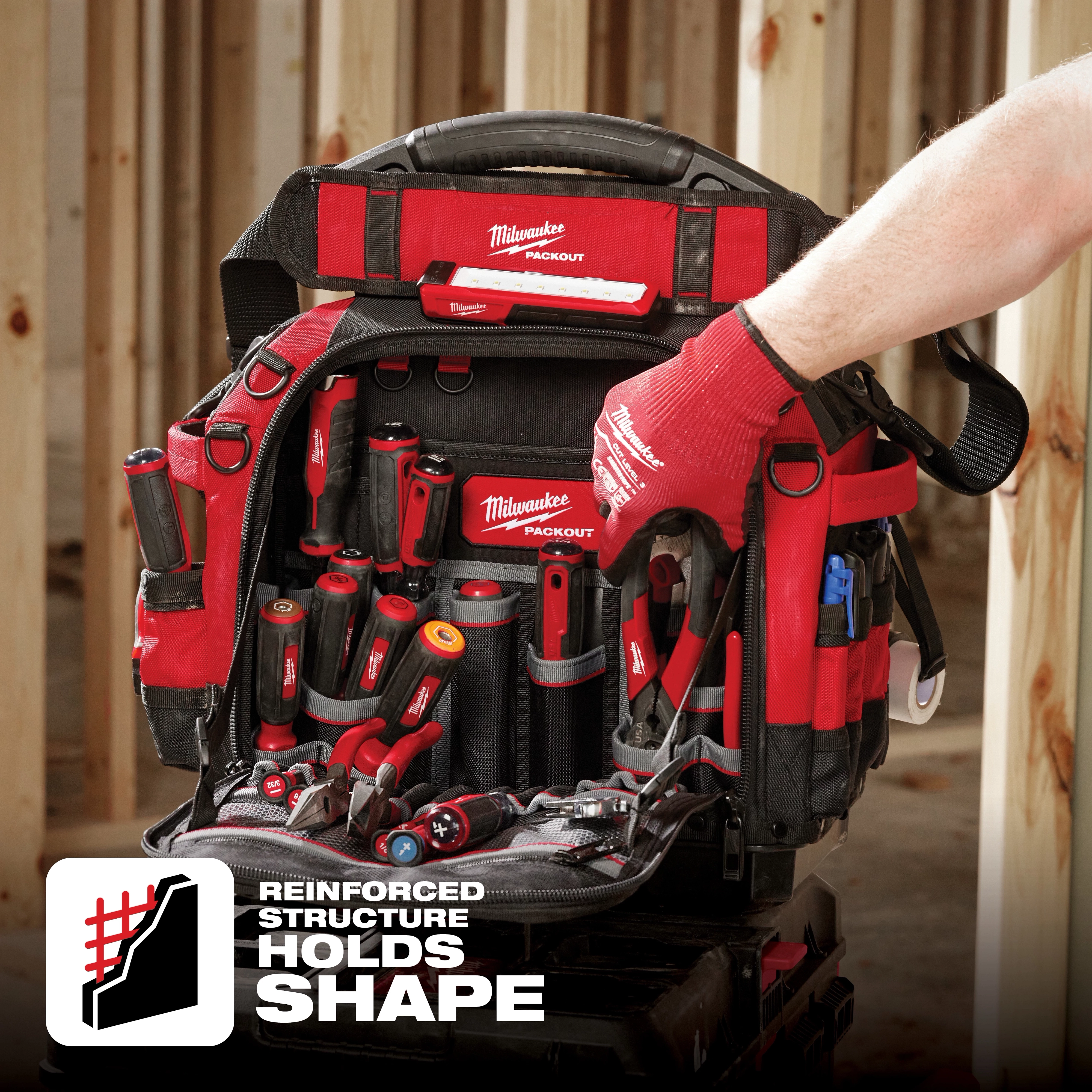 Milwaukee Packout tool bag with multiple tools inside. A person wearing red gloves is holding the handle. Text reads "Reinforced Structure Holds Shape".