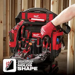 Milwaukee Packout tool bag with multiple tools inside. A person wearing red gloves is holding the handle. Text reads "Reinforced Structure Holds Shape".