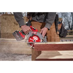 A person cuts wood using a circular saw equipped with a 7-1/4" 24T Thick Kerf Framing Circular Saw Blade. The saw is in use, generating wood dust. The person wears work clothes and a tool belt.