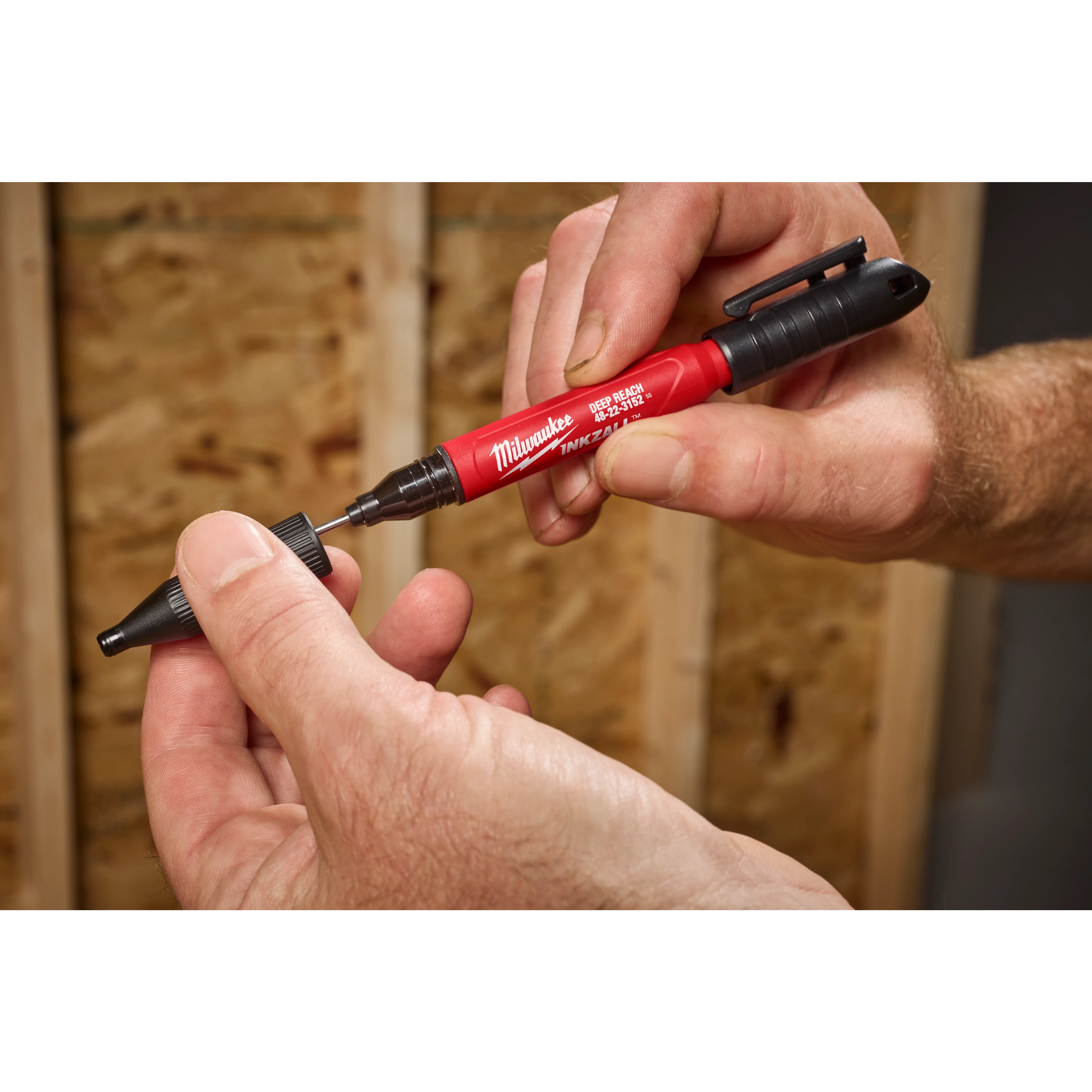 A person holds the INKZALL™ 2IN1 Deep Reach Ultra Fine Point Black Jobsite Marker in their right hand while removing the cap with their left hand. The marker is red with a black clip and tip, designed for precision marking on job sites.