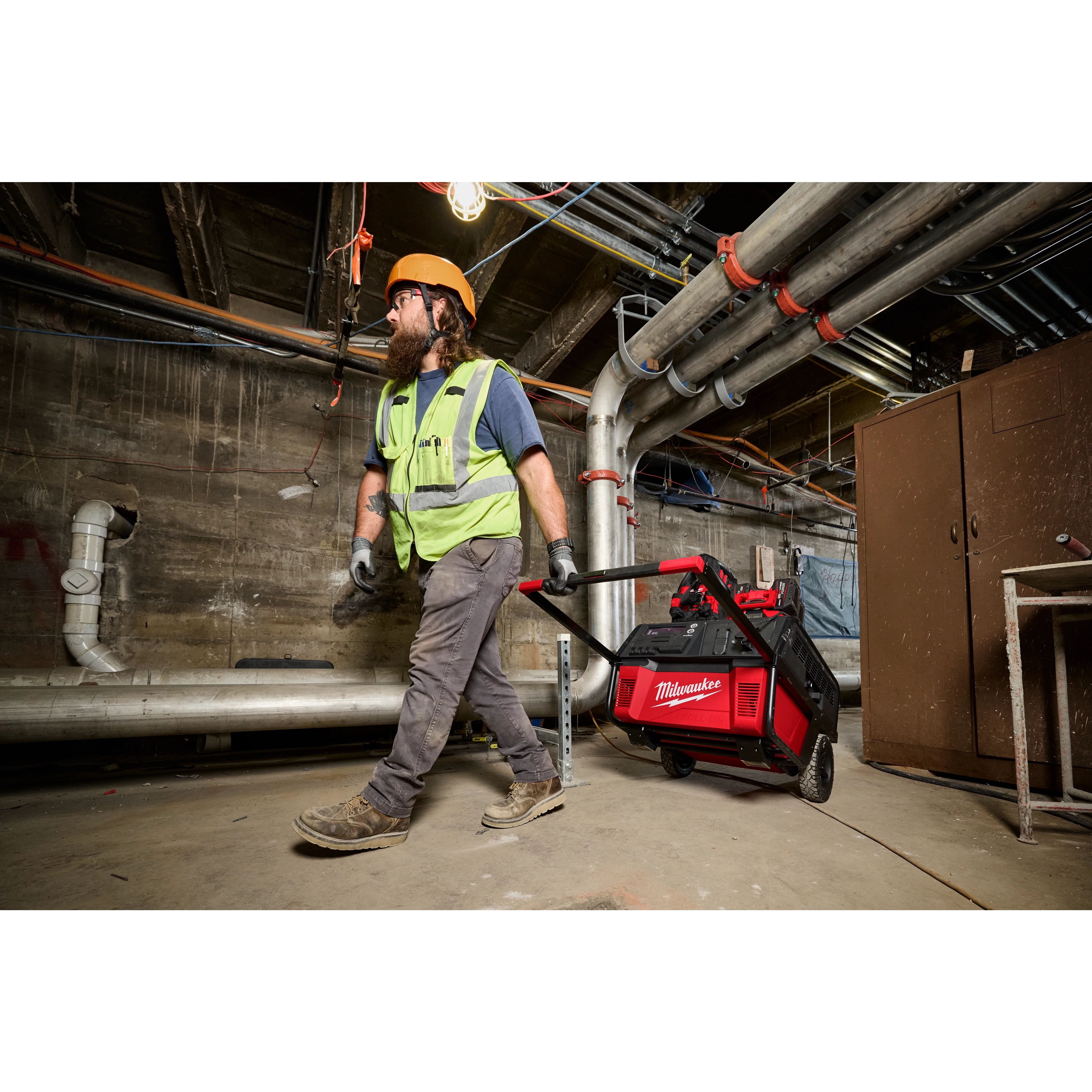 A construction worker wearing protective gear drags a red and black ROLL-ON™ 7200W/3600W 6.0kWh Power Supply through an industrial site. The power supply is mounted on a wheeled frame for easy transport. The background shows various pipes, cables, and concrete walls.