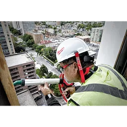 Worker on a high-rise building wears a white hard hat secured with a red Hard Hat Lanyard with Clip and holds a pipe.