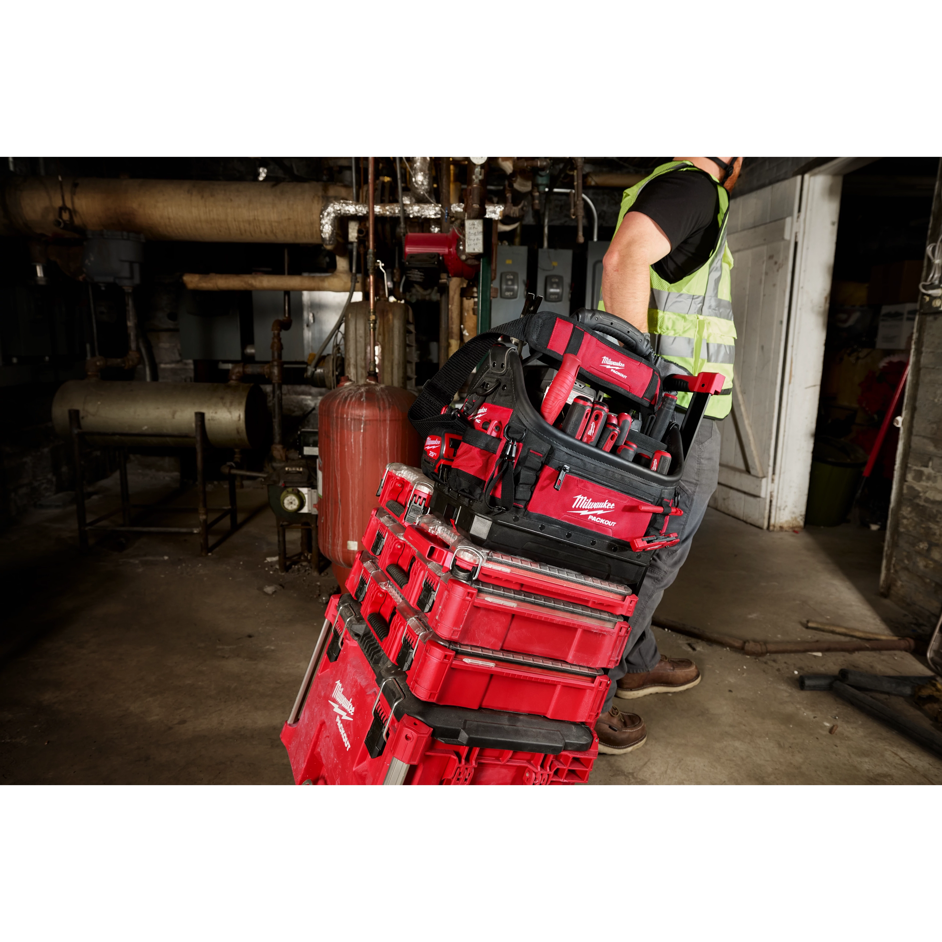 A worker in a high-visibility vest pulls a stack of red PACKOUT™ toolboxes, with the PACKOUT™ 15" Structured Tote on top. The tote is filled with various tools, and they are in an industrial setting with machinery and pipes in the background.