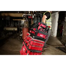 A worker in a high-visibility vest pulls a stack of red PACKOUT™ toolboxes, with the PACKOUT™ 15" Structured Tote on top. The tote is filled with various tools, and they are in an industrial setting with machinery and pipes in the background.