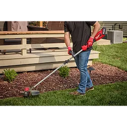 A person using the M18™ Brushless String Trimmer to trim grass along the edge of a garden bed. The trimmer has a red and black design with a long shaft and battery pack at the end. The user is wearing red gloves and jeans.