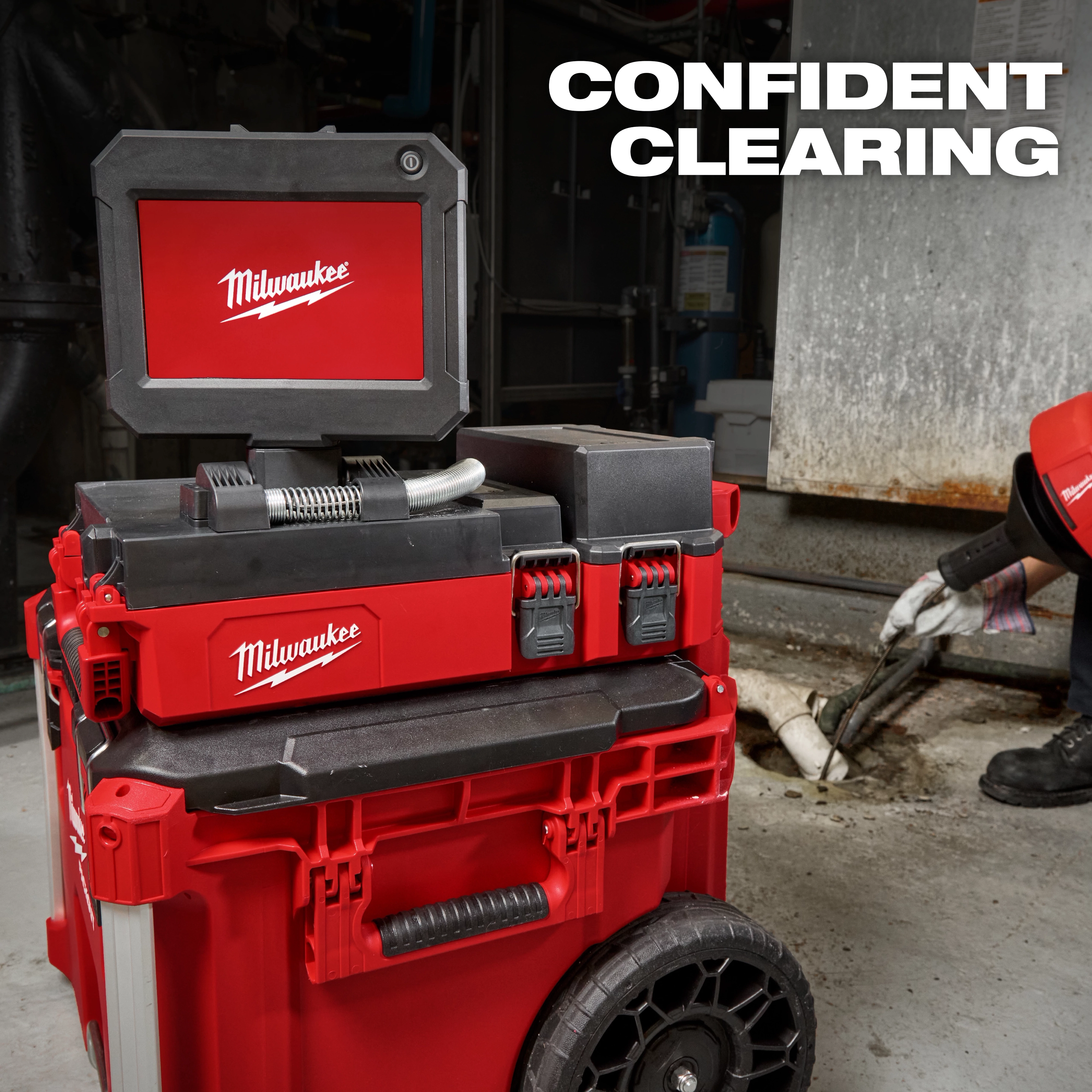 Image of the Milwaukee M12™ 75' Drain Camera with PACKOUT™ Compatibility in use by a worker. The red camera system features a screen and control unit mounted on a wheeled storage unit. Text "CONFIDENT CLEARING" is displayed in the top-right corner.