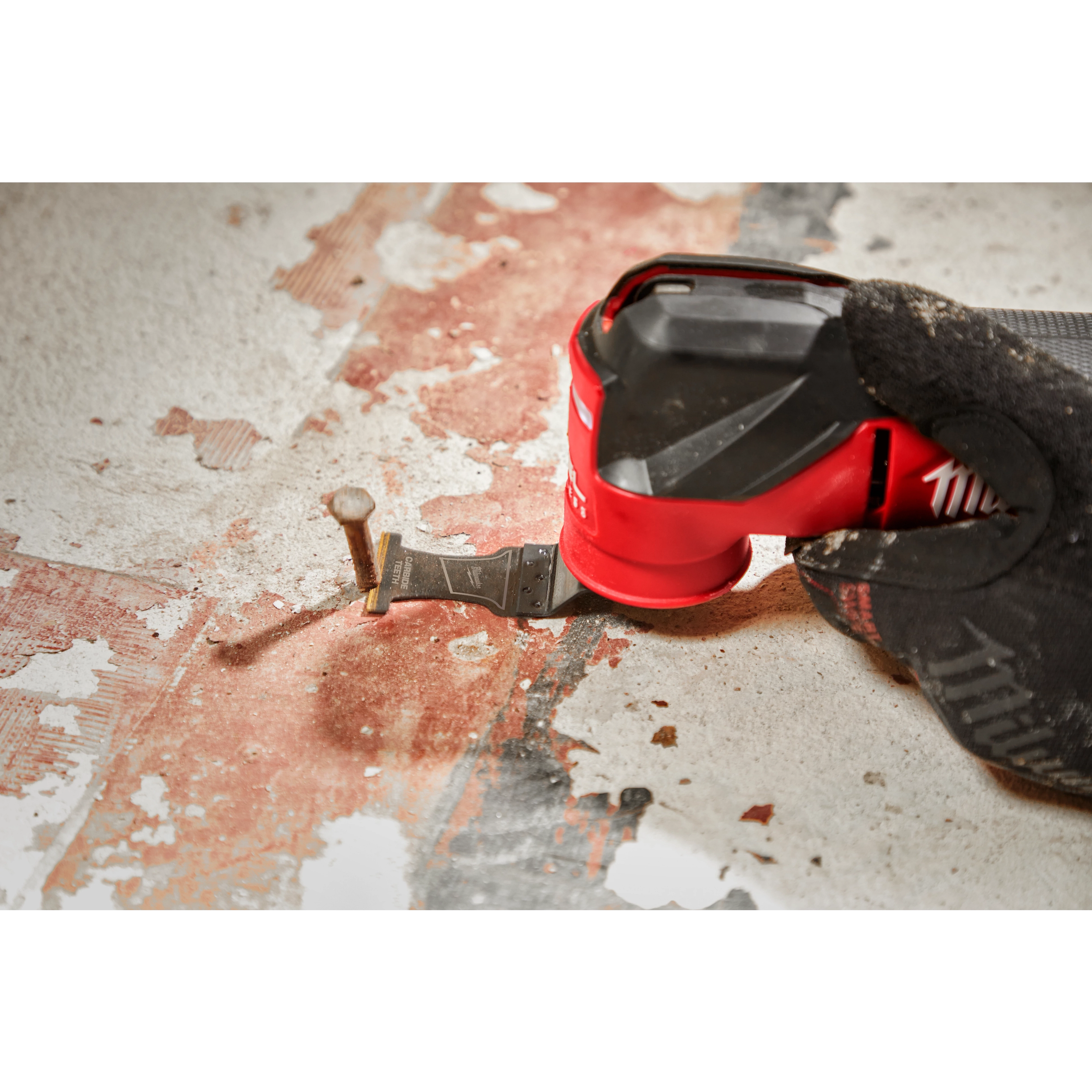A worker using a MILWAUKEE® OPEN-LOK™ 1-3/8" Carbide Teeth Metal Multi-Tool Blade to cut through an exposed nail on a worn floor. The hand and tool are focused in the foreground, demonstrating the blade's precision in cutting metal.