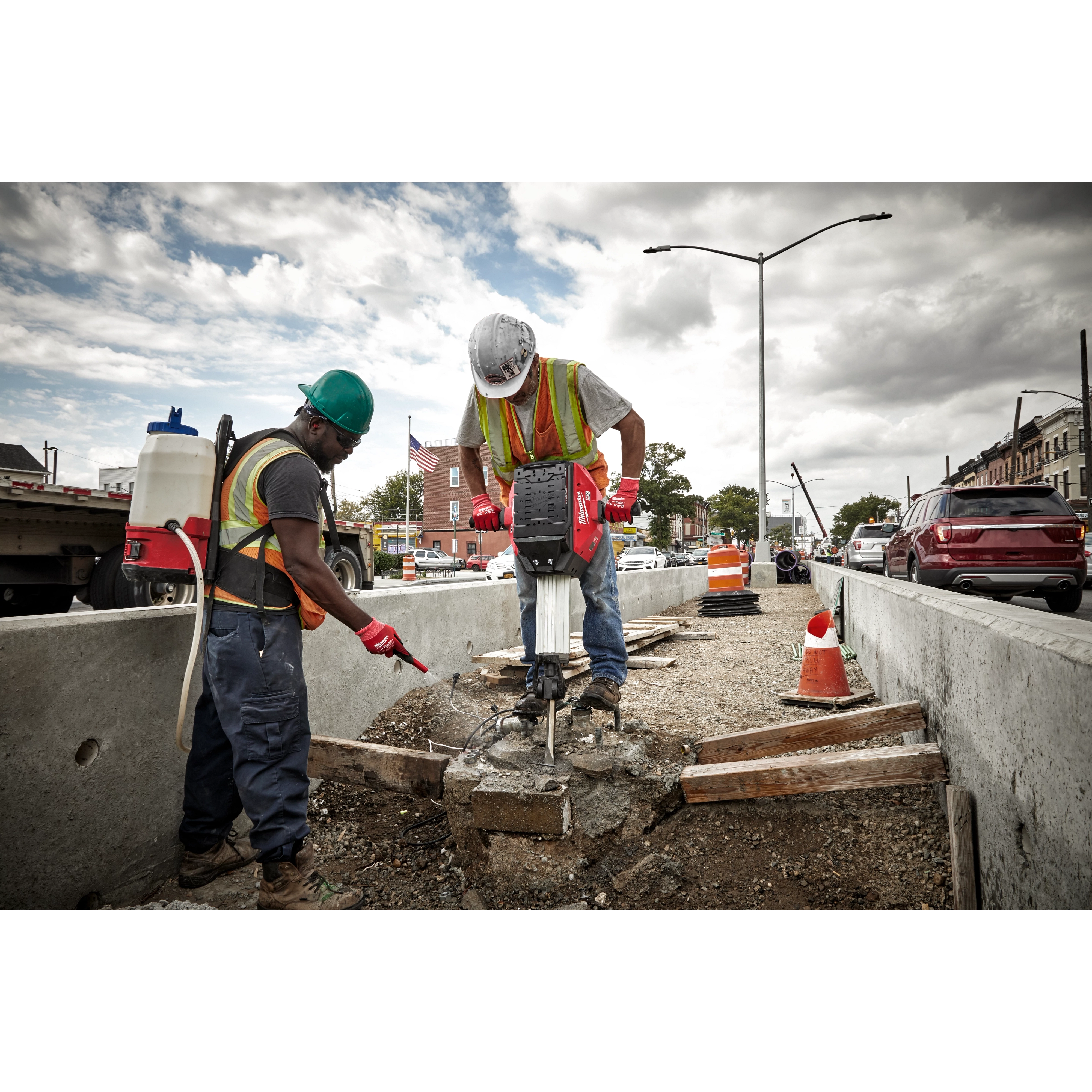 Two construction workers in safety gear use the MX FUEL™ Breaker to break up concrete on a roadside job site. One worker operates the breaker, while the other stands nearby pointing. Traffic cones and cars are visible in the background.