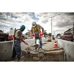 Two construction workers in safety gear use the MX FUEL™ Breaker to break up concrete on a roadside job site. One worker operates the breaker, while the other stands nearby pointing. Traffic cones and cars are visible in the background.