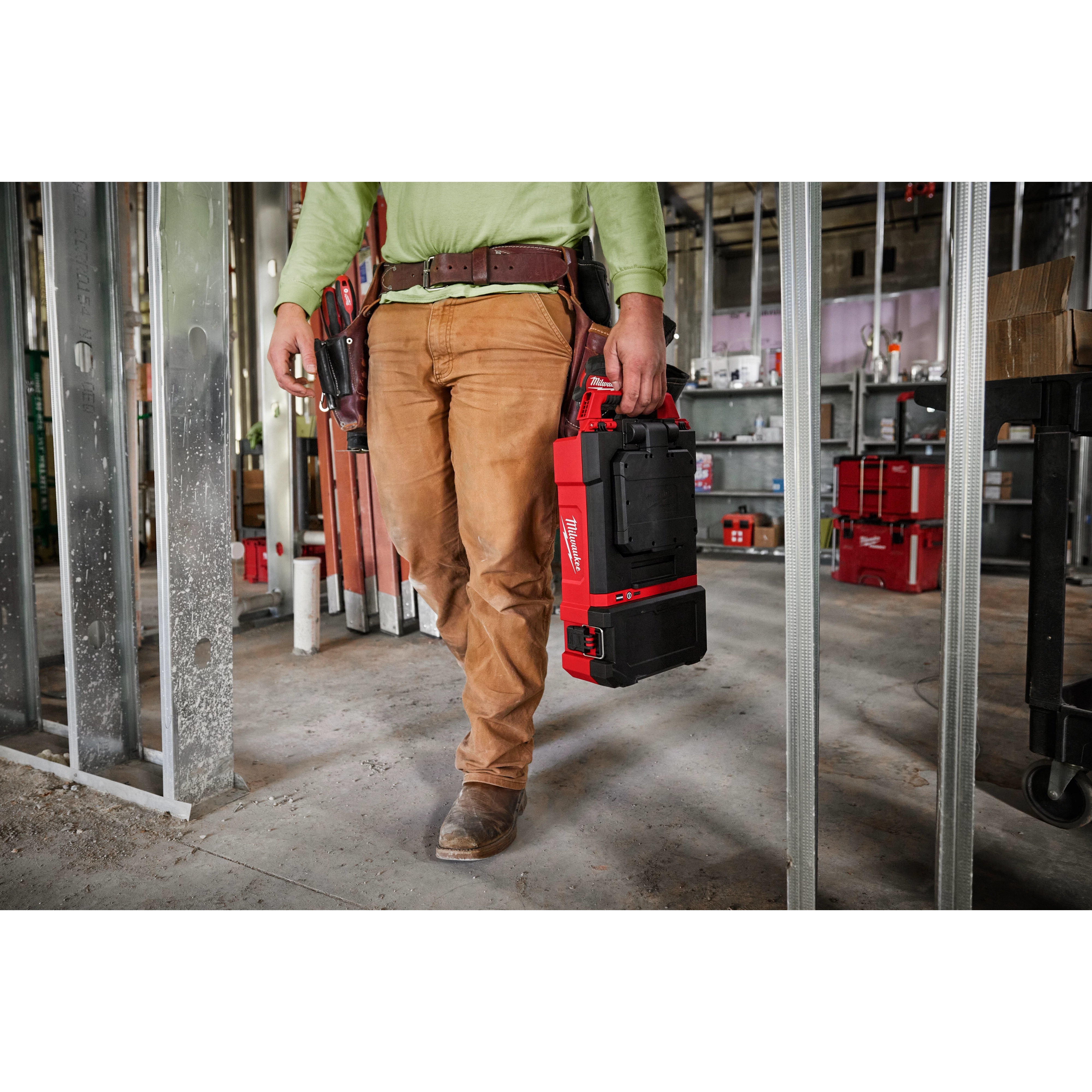 Worker carrying the M12 PACKOUT Flood Light with USB Charging in a construction site, with additional tools and equipment in the background.