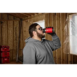 Person drinking from a PACKOUT 12oz Insulated Bottle with Sip Lid in a construction environment. Red tool storage boxes in the background.