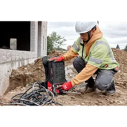 A person in construction gear uses the MX FUEL™ Portable Pump Power Base and MX FUEL™ 1HP 2" Submersible Pump Kit at a construction site. The equipment is placed on the ground with cables lying around it. The environment includes dirt and a partially constructed building.