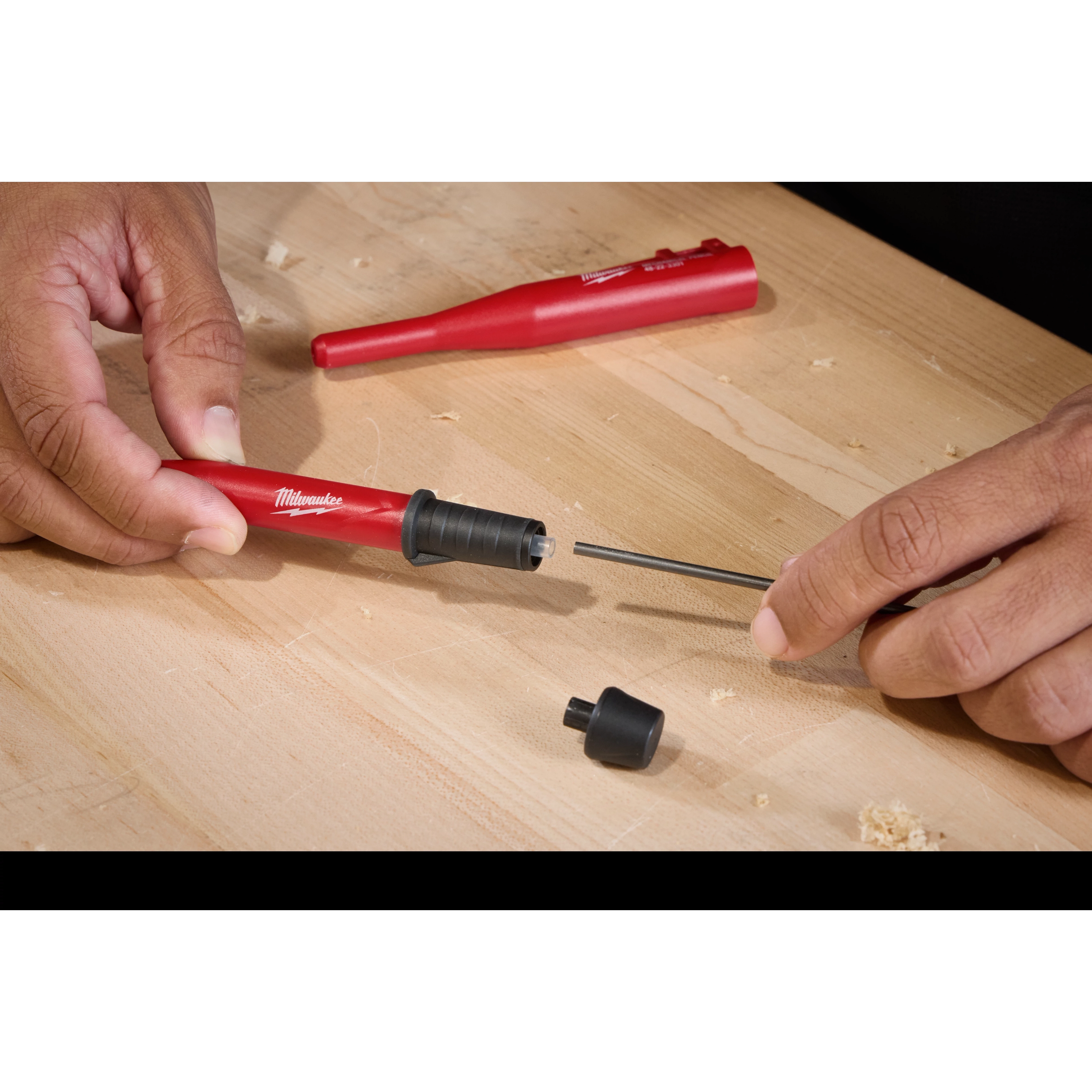 Two hands assemble a Milwaukee Mechanical Pencil on a light wooden surface. The pencil is being prepared with a black lead core. A small black cap is also visible on the surface.