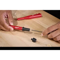 Two hands assemble a Milwaukee Mechanical Pencil on a light wooden surface. The pencil is being prepared with a black lead core. A small black cap is also visible on the surface.
