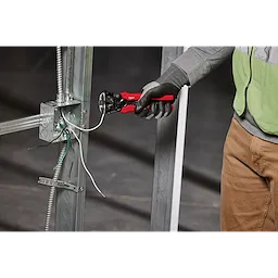 A person wearing gloves and a safety vest uses a Self-Adjusting Wire Stripper & Cutter to strip a wire. The tool has red handles and is positioned next to a metallic electrical panel.