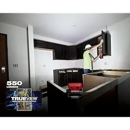 A person using a REDLITHIUM USB ROVER Pivoting Flood Light to illuminate a partially constructed kitchen with dark wood cabinets.