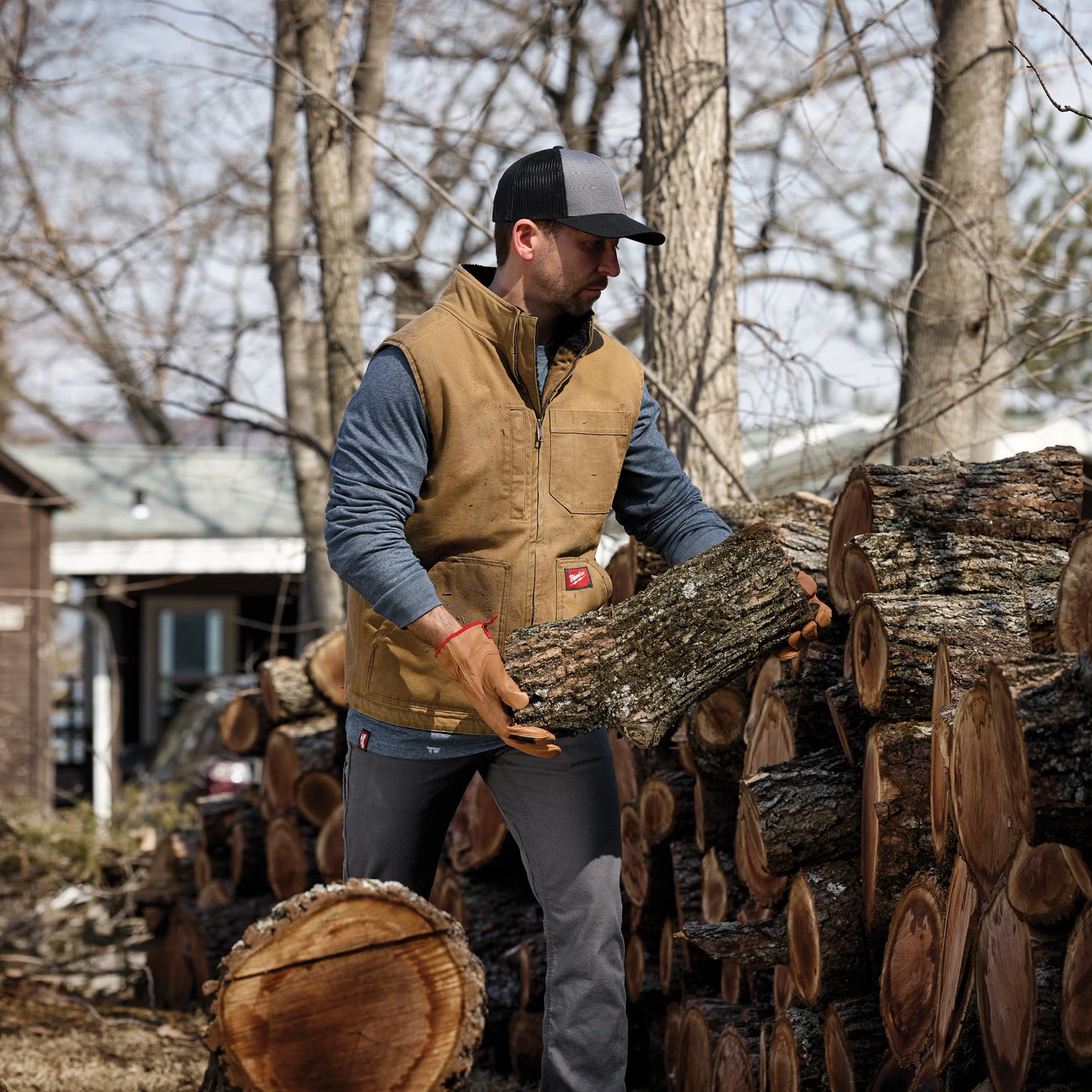 Man stacking firewood while wearing a tan vest, gray sleeves, gloves, and GRIDIRON Snapback Trucker Hat - Gray in a wooded area.