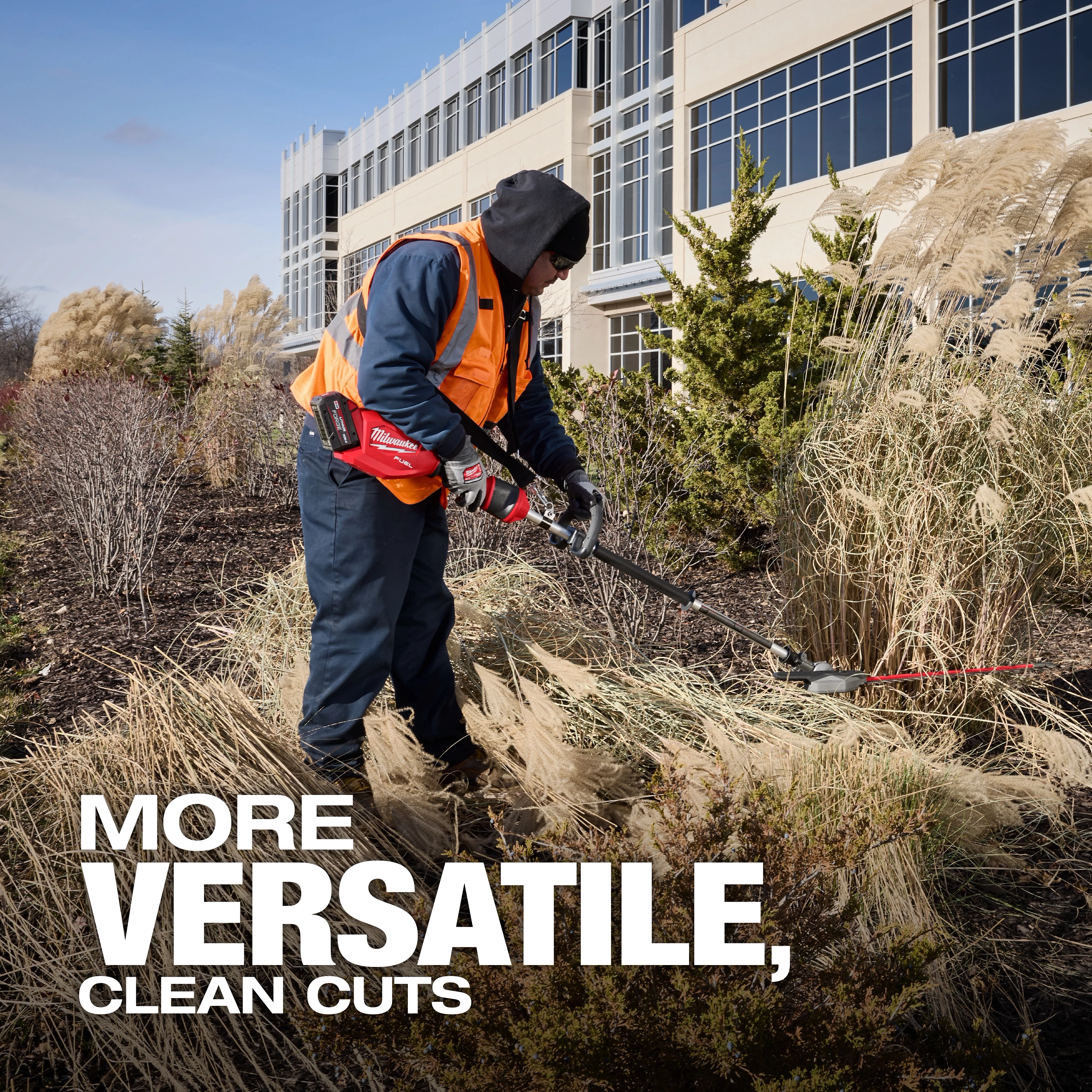 A worker trims tall grass with a power trimmer in front of a large office building. The image includes the text, "MORE VERSATILE, CLEAN CUTS" at the bottom. The worker wears a high-visibility vest and gloves.