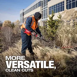 A worker trims tall grass with a power trimmer in front of a large office building. The image includes the text, "MORE VERSATILE, CLEAN CUTS" at the bottom. The worker wears a high-visibility vest and gloves.