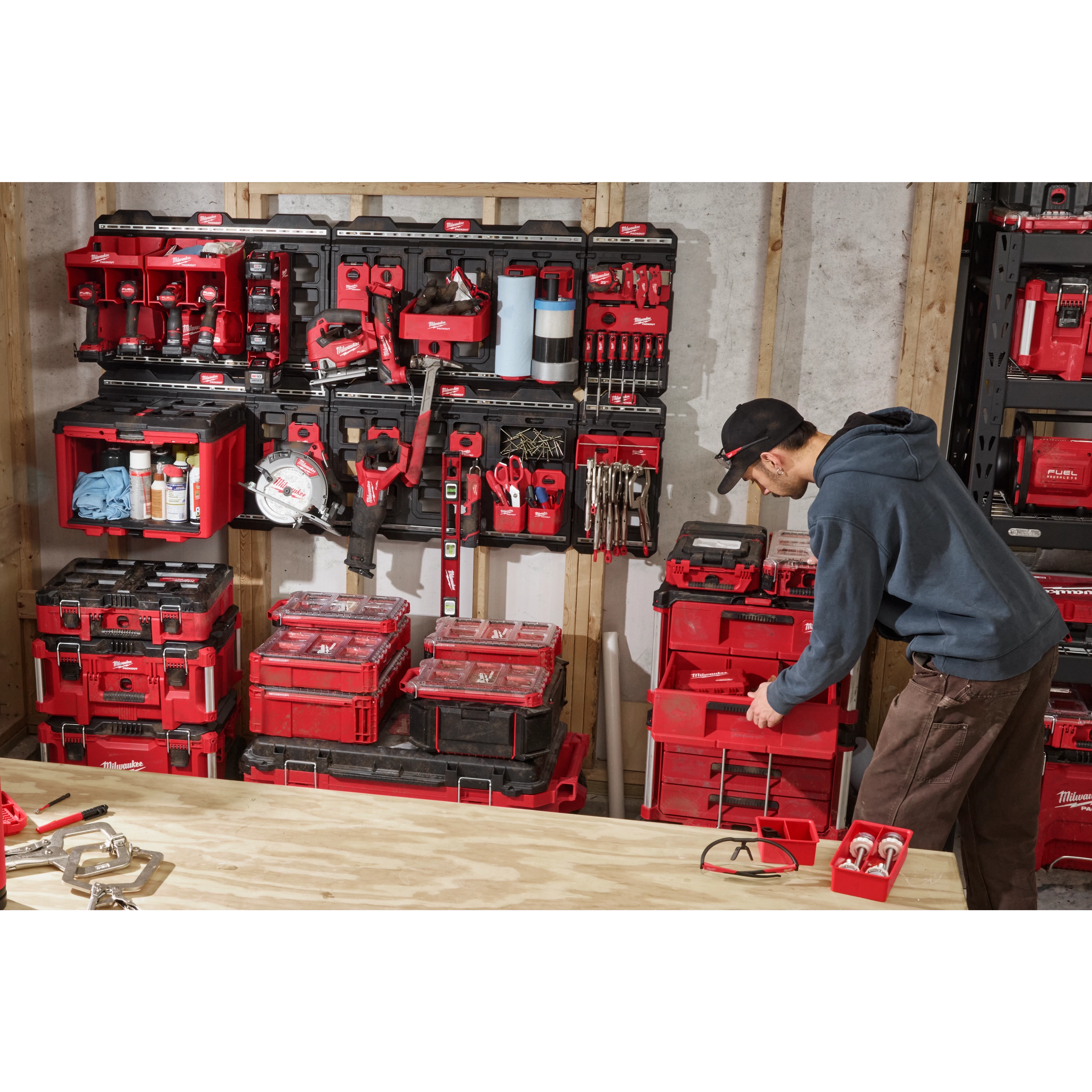 A man organizes various red and black tool storage boxes and organizer units on a PACKOUT™ Large Wall Plate mounted on a wooden frame. The workspace is filled with tools and accessories arranged neatly on multiple shelves, creating an efficient and organized tool storage area.