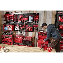 A man organizes various red and black tool storage boxes and organizer units on a PACKOUT™ Large Wall Plate mounted on a wooden frame. The workspace is filled with tools and accessories arranged neatly on multiple shelves, creating an efficient and organized tool storage area.