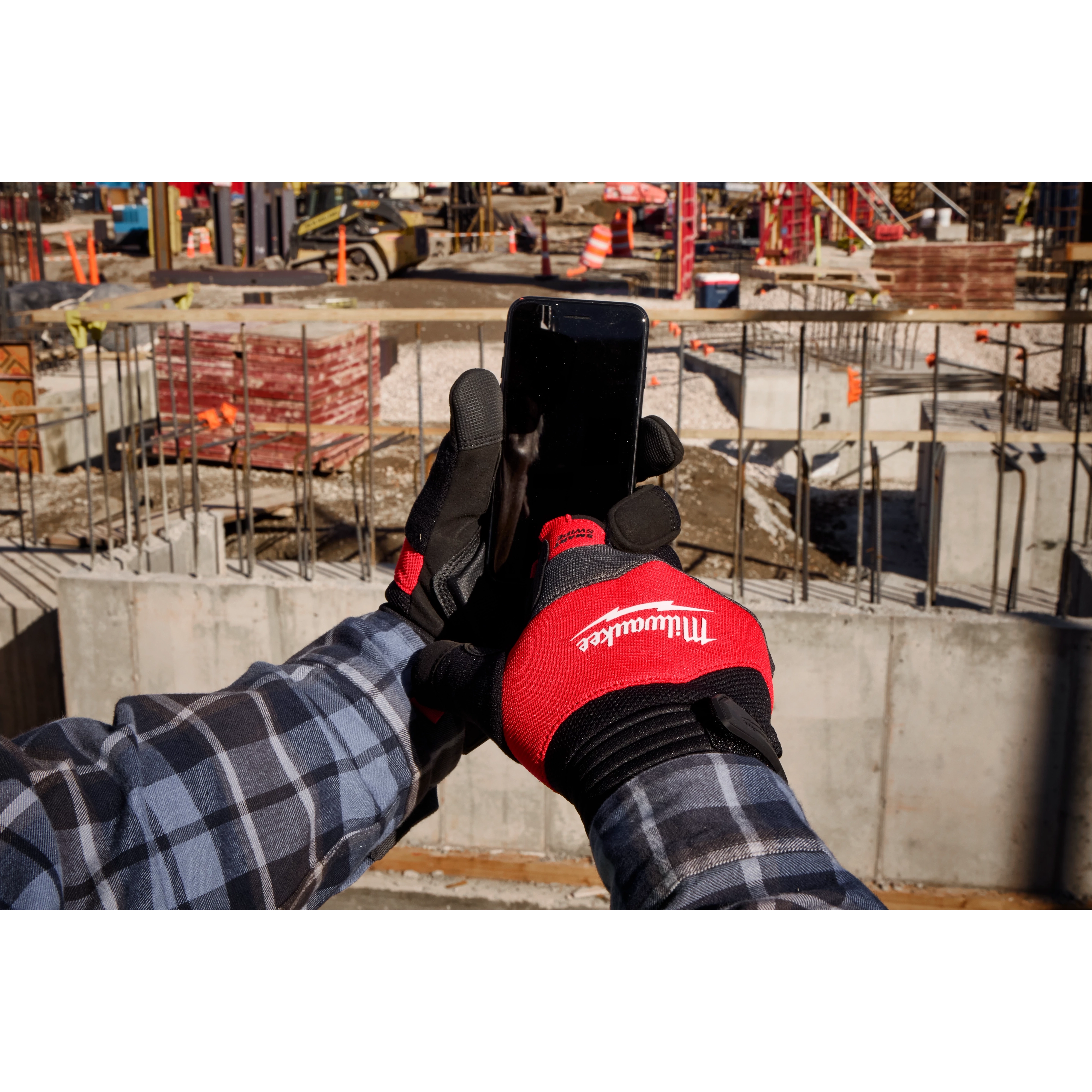 A person wearing a red and black Anti-Vibration Work Glove is holding a smartphone at a construction site. The gloves have the logo of "Milwaukee" on them. The background features construction materials, equipment, and unfinished structures.