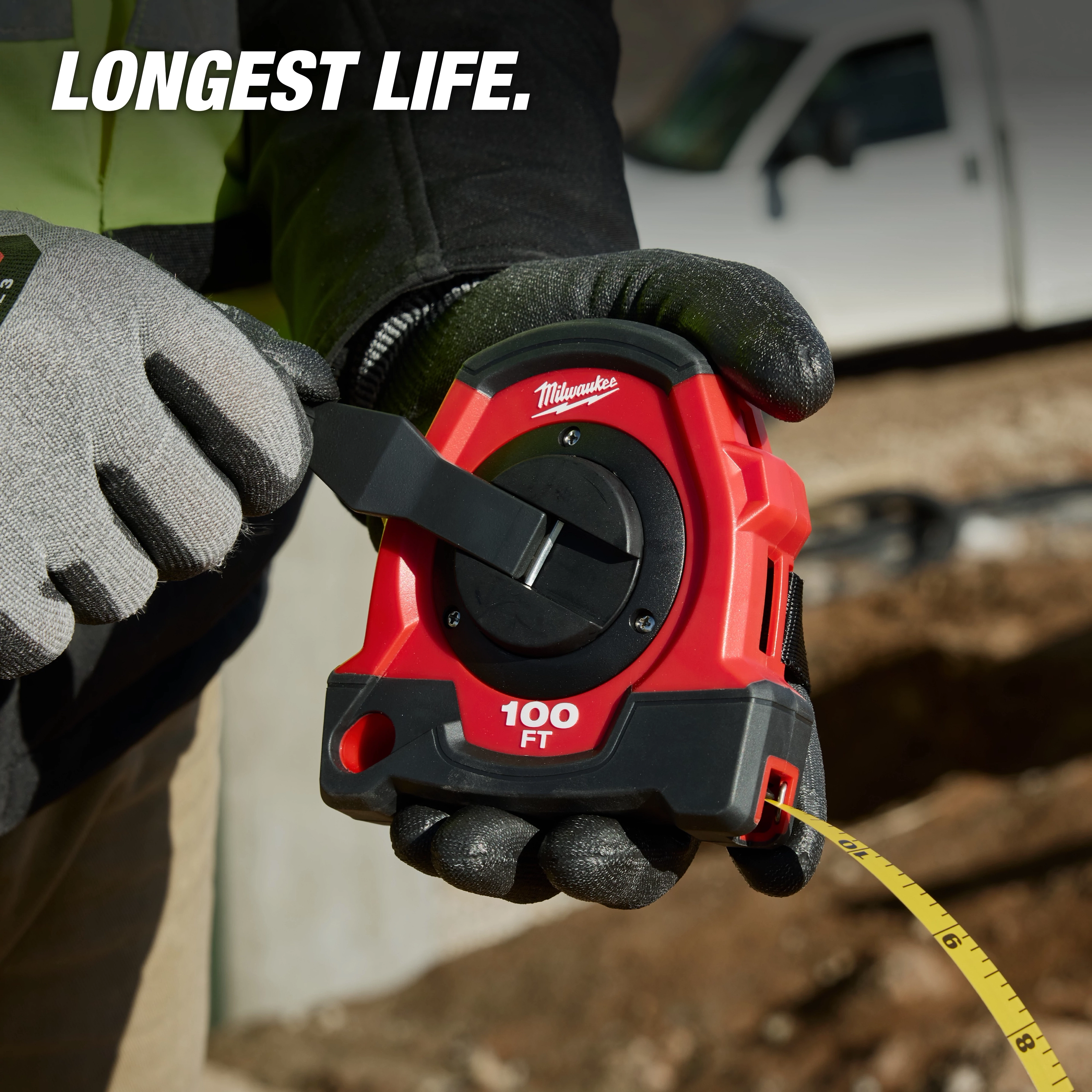 Gloved hands holding a red 100FT Closed Reel Long Tape with extended yellow tape at a construction site.