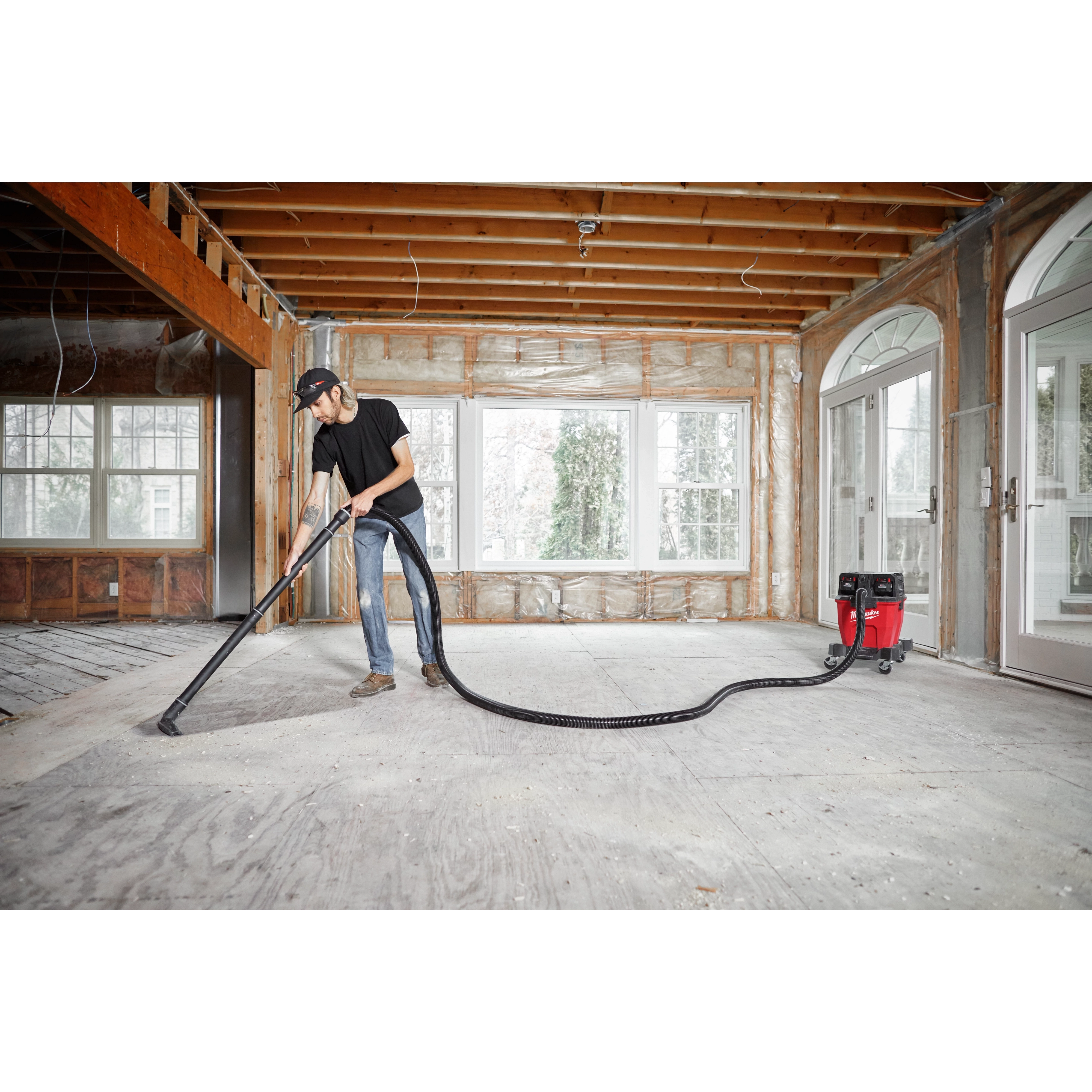 Man using a vacuum cleaner attached to a 1-7/8” x 16' flexible hose in an unfinished room with exposed insulation and large windows.