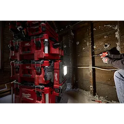 A stack of red Milwaukee PACKOUT™ Tool Boxes, including the PACKOUT™ Tool Box Compact Plate, is positioned against a wall with exposed studs. A person on the right uses a power drill on the wall, and a bright work light from the toolboxes illuminates the scene.