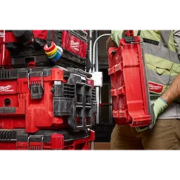 The image shows a worker holding a red PACKOUT™ Tool Box Compact Plate alongside a stack of other red and black PACKOUT™ toolboxes. The compact plate has multiple latches and a handle for portability, and is part of a modular storage system. The worker is wearing gloves and a green safety vest, indicating the setting might be a construction or industrial environment.