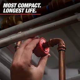 A hand is adjusting a red Liberty Pump check valve installed in a copper pipe system. The accompanying text reads "Most Compact. Longest Life." indicating the product’s features.