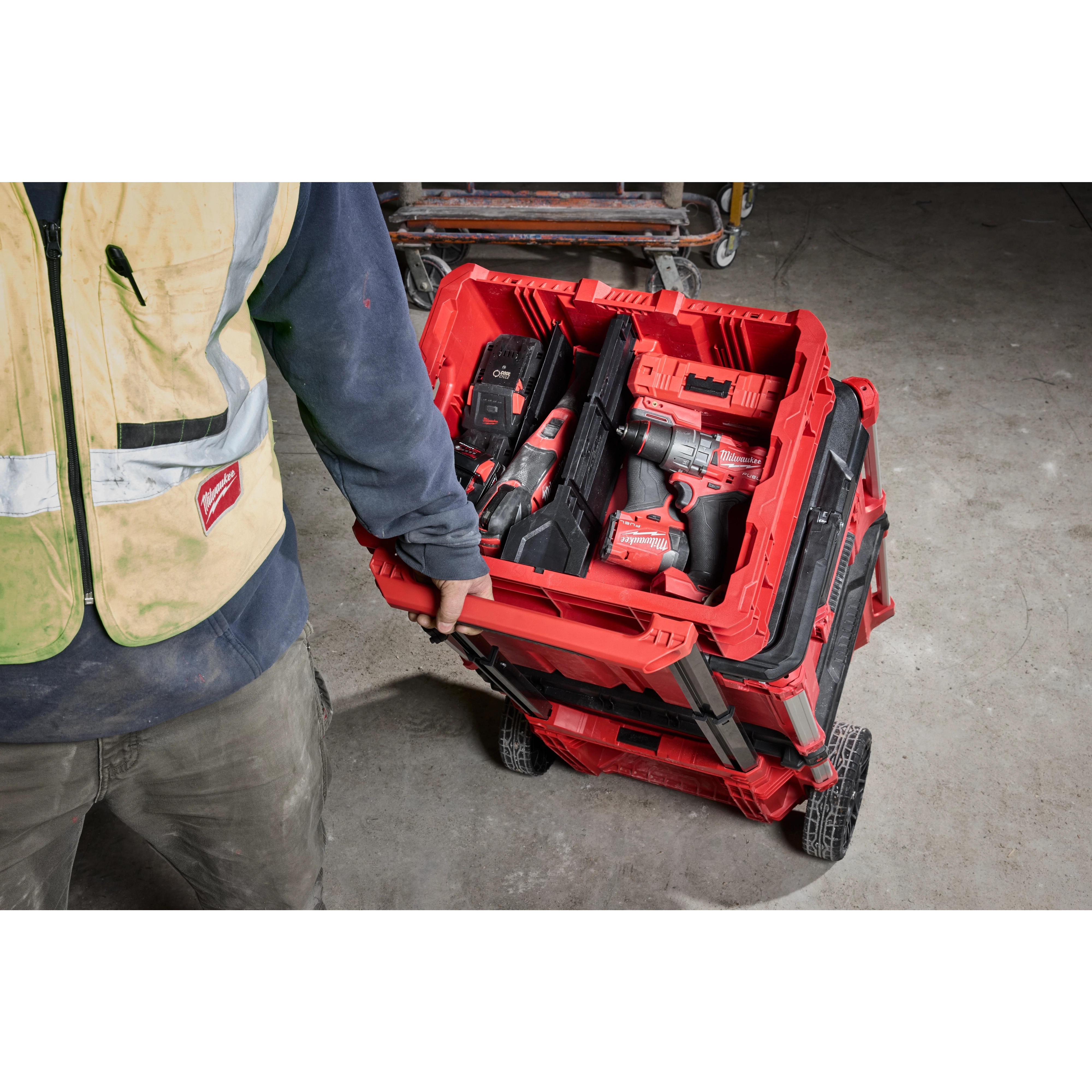 A person in a safety vest is pulling a loaded PACKOUT Low-Profile Crate on a wheeled cart over a concrete floor.