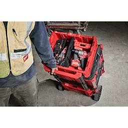 A person in a safety vest is pulling a loaded PACKOUT Low-Profile Crate on a wheeled cart over a concrete floor.