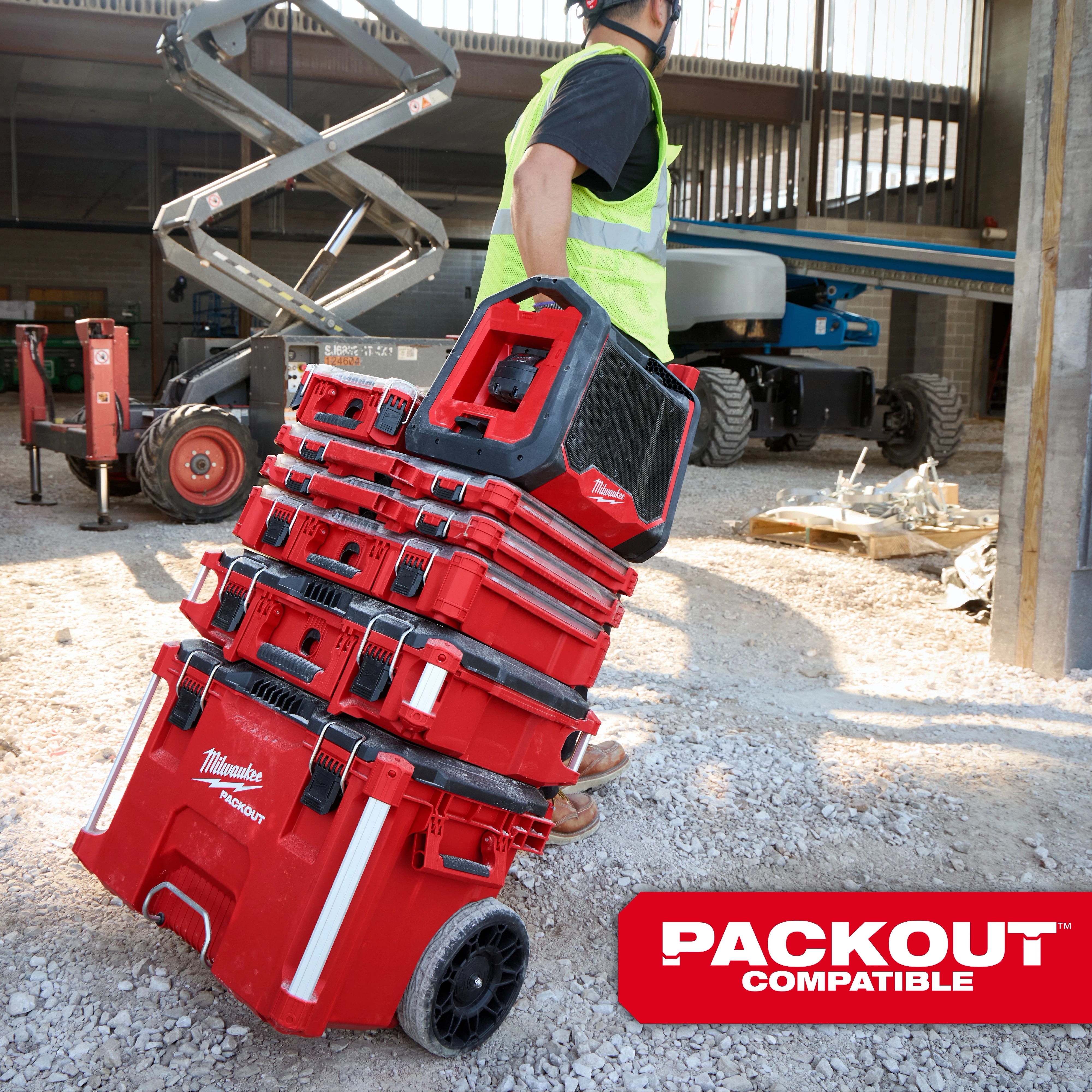 Person hauling stacked Milwaukee Packout storage boxes on a construction site. The M18 BLUETOOTH JOBSITE RADIO + CHARGER is seen on top of the stack.