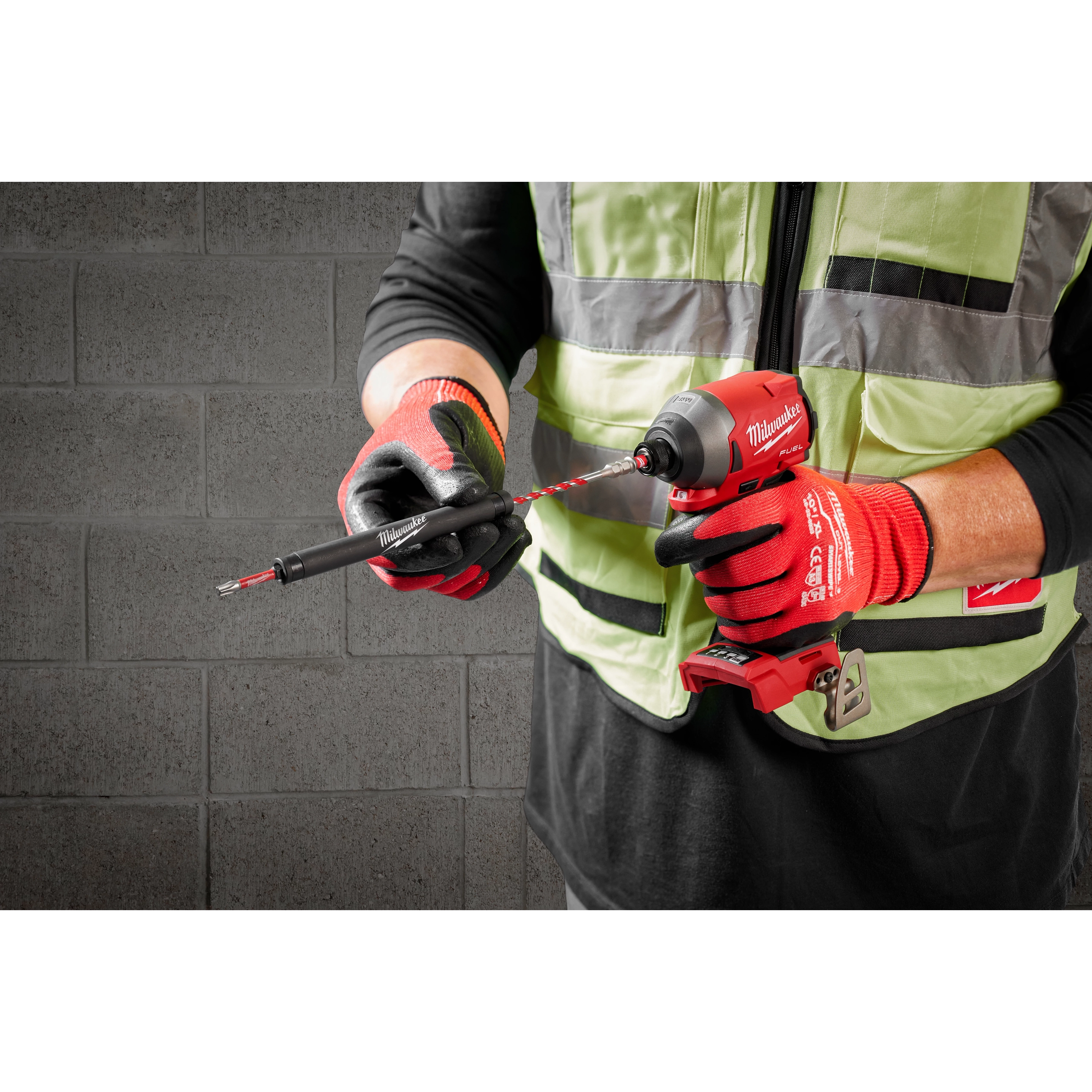 A person wearing a black shirt, green safety vest, and red gloves uses a red handheld drill to attach a drill bit. The drill bit is part of the 7pc. SHOCKWAVE Impact Duty™ Carbide Hammer Drill Bit Concrete Screw Install Kit, suitable for drilling into concrete.