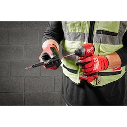 A person wearing a black shirt, green safety vest, and red gloves uses a red handheld drill to attach a drill bit. The drill bit is part of the 7pc. SHOCKWAVE Impact Duty™ Carbide Hammer Drill Bit Concrete Screw Install Kit, suitable for drilling into concrete.