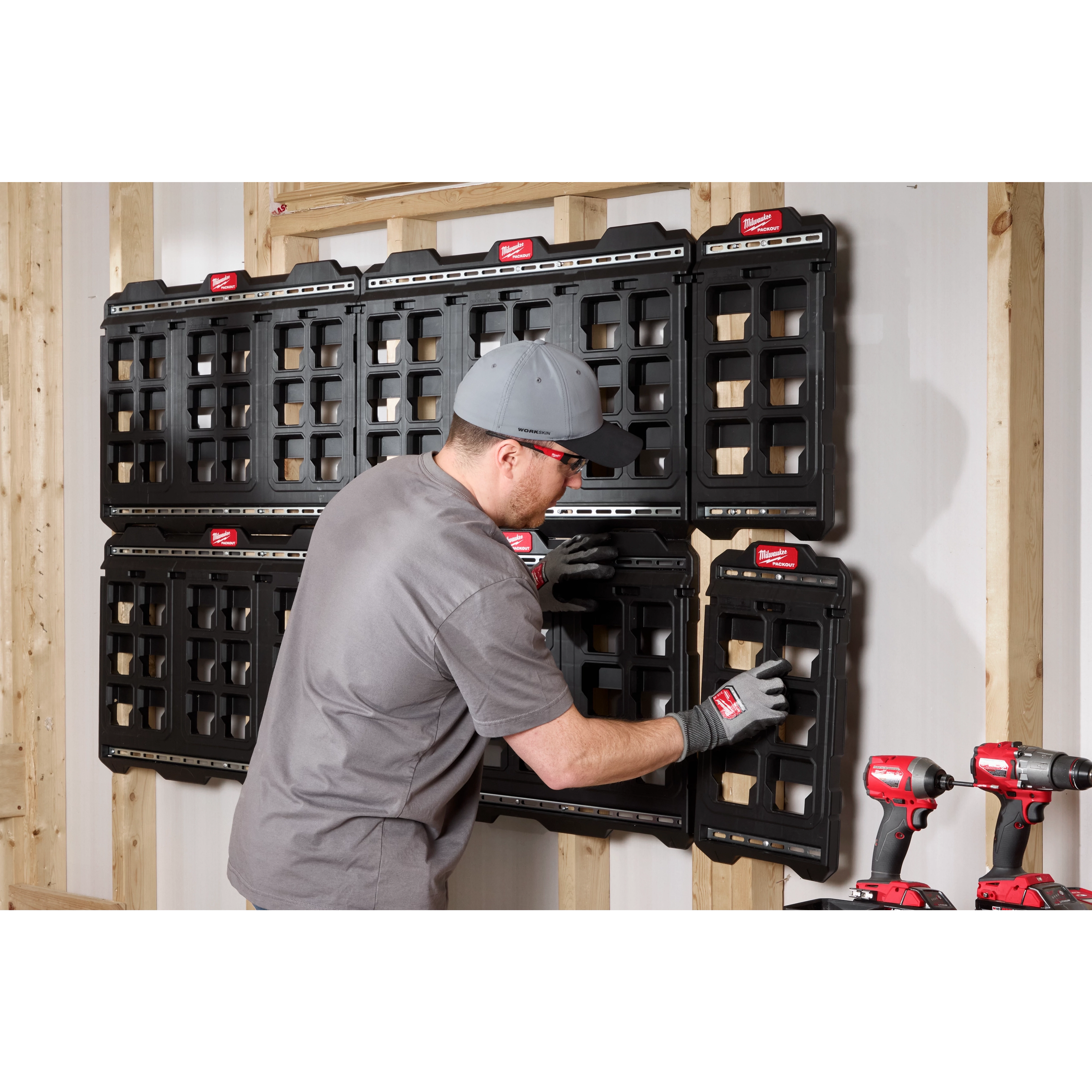 A person is mounting a PACKOUT™ Large Wall Plate on a wooden wall. Several black wall plates are already installed. Two cordless drills and a battery charger are positioned at the lower right side of the image. Other wall plates remain empty, waiting to be filled with tools.