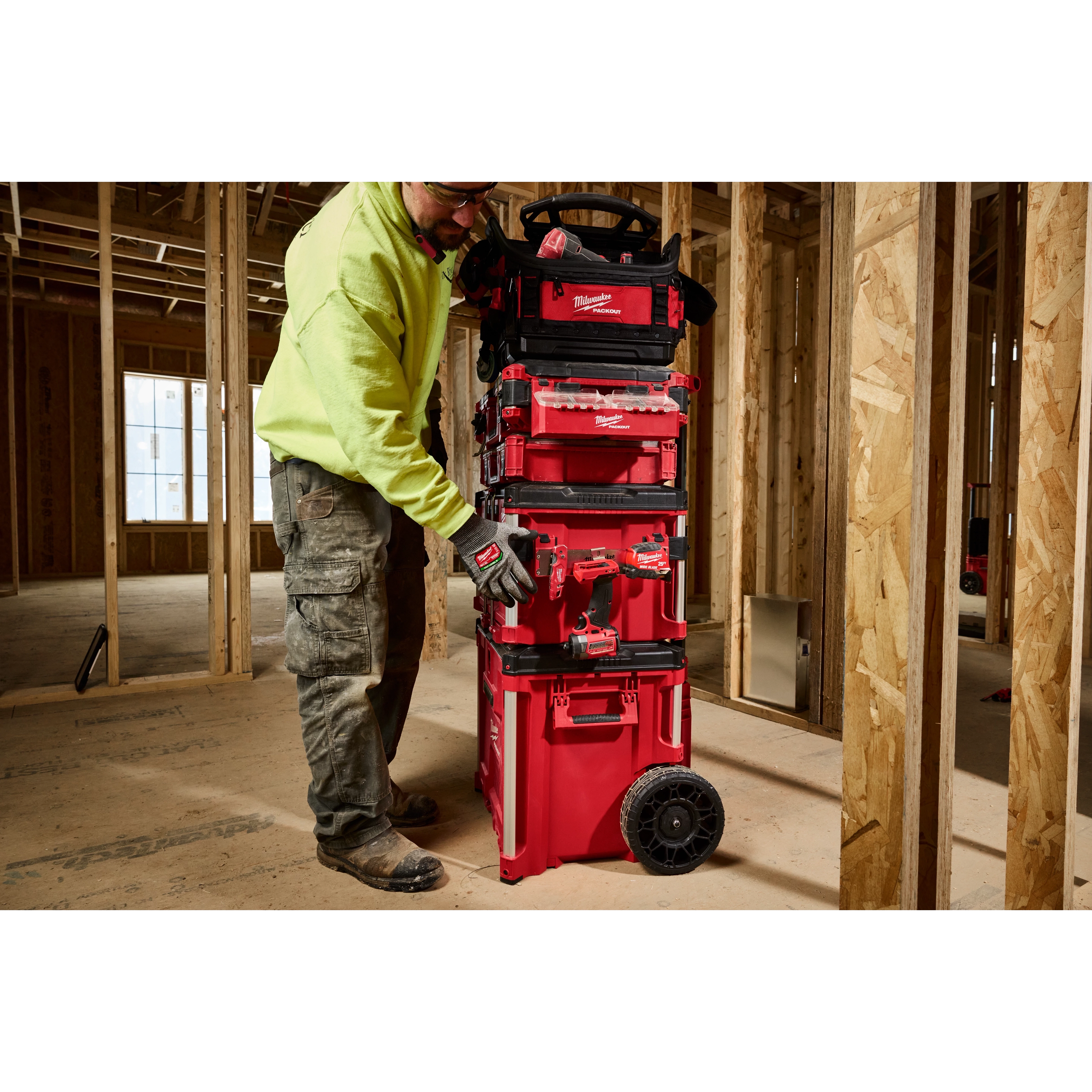 A person in a construction site interacts with a red PACKOUT Tool Box Belt Clip Rack Attachment, which is stacked with other toolboxes.