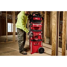 A person in a construction site interacts with a red PACKOUT Tool Box Belt Clip Rack Attachment, which is stacked with other toolboxes.