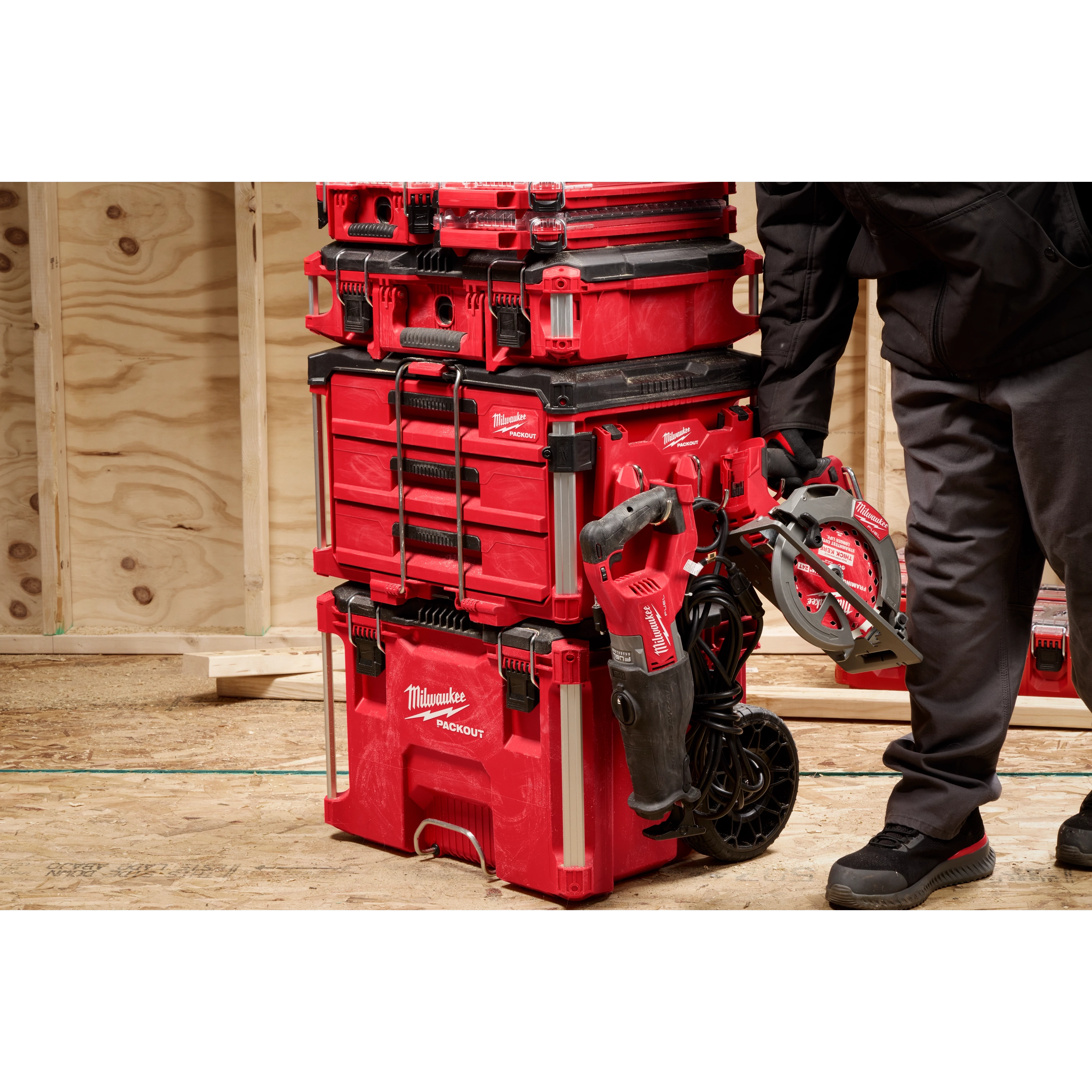 Person placing a red power tool next to a stack of red Milwaukee toolboxes on a wooden floor.