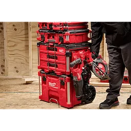 Person placing a red power tool next to a stack of red Milwaukee toolboxes on a wooden floor.