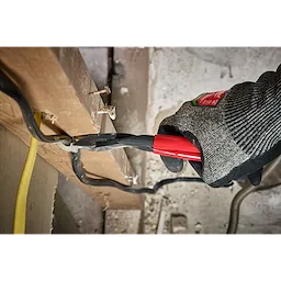 A gloved hand using Knipex High Leverage Diagonal Cutters cuts through a black wire attached to a wooden beam. The tool features red handles and is being used in a construction or renovation setting.