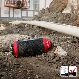The image shows an M12™ Bluetooth® Jobsite Speaker w/ PACKOUT™ Compatibility lying on a rugged, outdoor construction site. The speaker is black with red accents and is positioned among dirt, rocks, and a pipe. Icons indicate its IP64 dust and water resistance, as well as impact resistance.