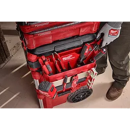 The image shows the PACKOUT™ Tool Box Caddy Attachment connected to a stack of red Milwaukee PACKOUT™ toolboxes. The caddy holds various Milwaukee tools, such as screwdrivers and pliers, and includes organization slots and pockets. A person wearing gloves is reaching for a screwdriver.