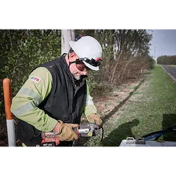 A worker wearing a BOLT™ White Full Brim Safety Helmet – Type 2, Class E, uses an electric drill outdoors. The worker is dressed in a green and black jacket, brown gloves, and is standing next to a vehicle on the side of a rural road with trees in the background.