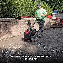 A person uses the MX FUEL™ 20" Plate Compactor Kit on a dirt pathway next to a brick wall and bushes. The compactor has a black and red body with the brand name visible. Various tools and equipment are also visible in the background.