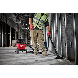 A worker in a green safety vest and red gloves uses a Milwaukee M18 FUEL 6 Gallon Wet/Dry Vacuum to clean a construction site.
