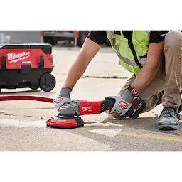 A worker uses the Milwaukee 7"/9" Surface Grinding Dust Shroud attached to a grinding tool on a concrete surface. The dust extraction vacuum is connected for debris collection.