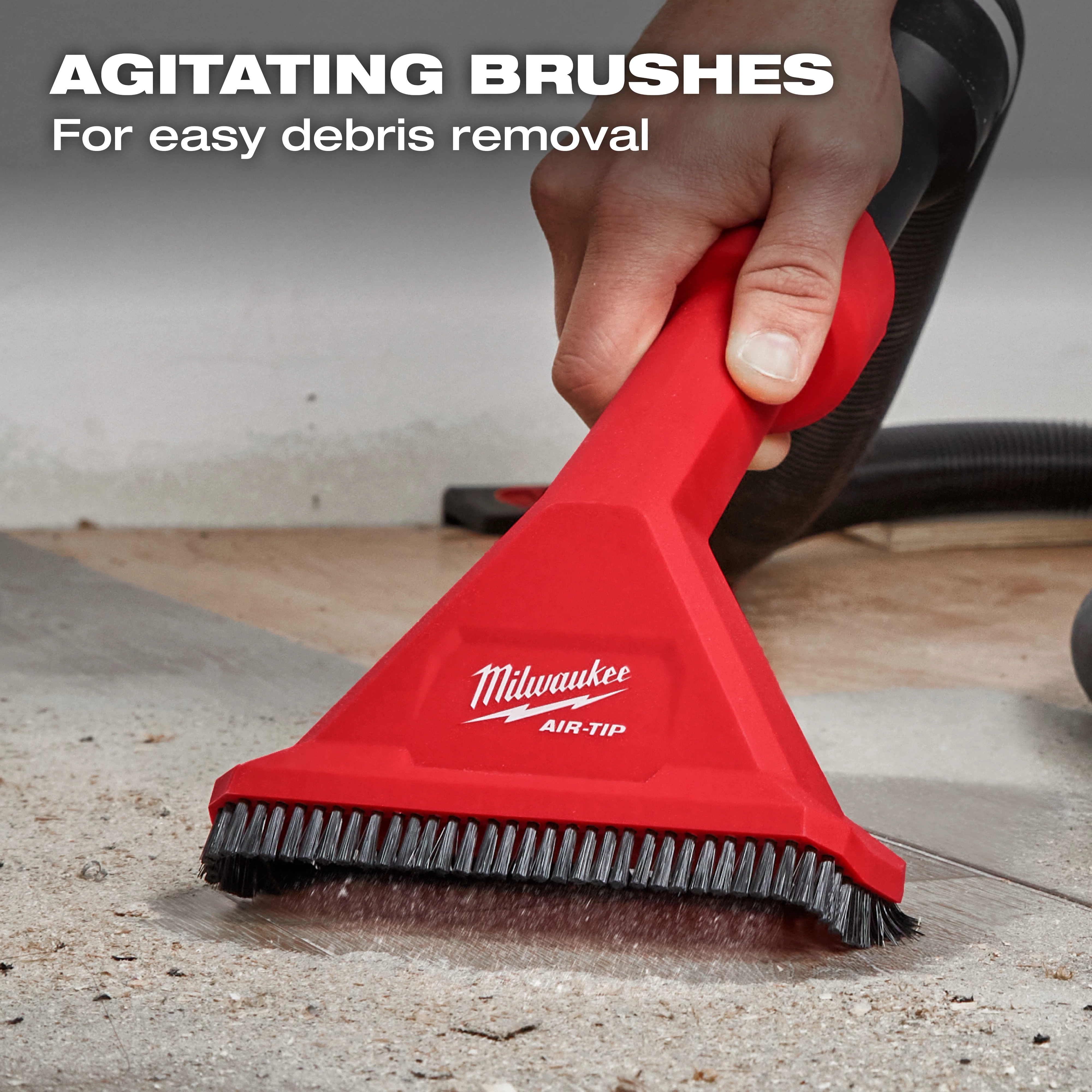 A hand uses the Milwaukee AIR-TIP™ Rocking Utility Nozzle w/ Brushes on a dirty surface. The nozzle is red with black bristles and labeled "AIR-TIP™." In the background, text reads "AGITATING BRUSHES For easy debris removal."